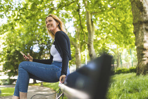 Lächelnde reife Frau mit Smartphone schaut weg, während sie auf einer Parkbank sitzt - WPEF04621