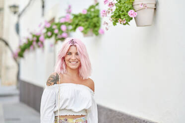 Lächelnde junge Frau mit rosa Haaren an der Wand stehend - KIJF03930