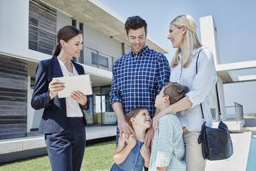 Businesswoman with digital tablet smiling while looking at family in front of villa - RORF02719