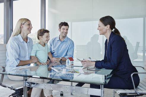 Glückliche Familie im Gespräch mit einer Immobilienmaklerin am Tisch sitzend - RORF02744