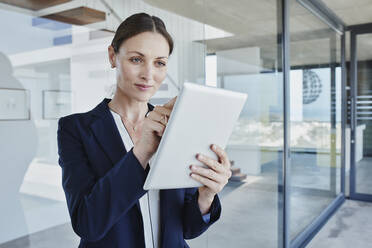 Geschäftsfrau, die ein digitales Tablet benutzt, während sie an einer Glaswand steht - RORF02754