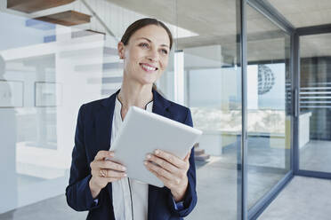 Beautiful smiling saleswoman with digital tablet standing by glass wall looking away - RORF02755