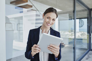 Smiling female real estate agent with digital tablet standing by glass wall - RORF02756