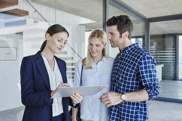 Mature businesswoman writing on clipboard while discussing with couple standing by glass wall - RORF02757