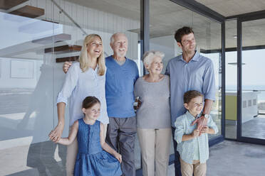 Smiling multi-generation family standing together in front of glass wall looking away - RORF02786