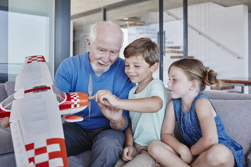 Ein älterer Mann hält ein Spielzeugflugzeug in der Hand, während er mit seinen Enkelkindern zu Hause im Wohnzimmer sitzt - RORF02788