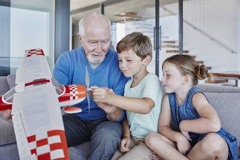 Großvater hält Spielzeugflugzeug, während er mit seinen Enkelkindern auf dem Sofa im Wohnzimmer sitzt - RORF02789