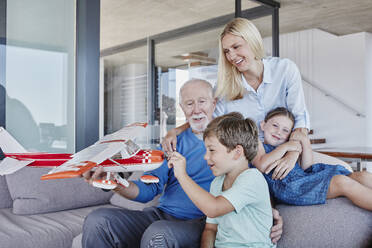 Senior man holding toy airplane playing with boy while sitting by daughter and girl in living room - RORF02790