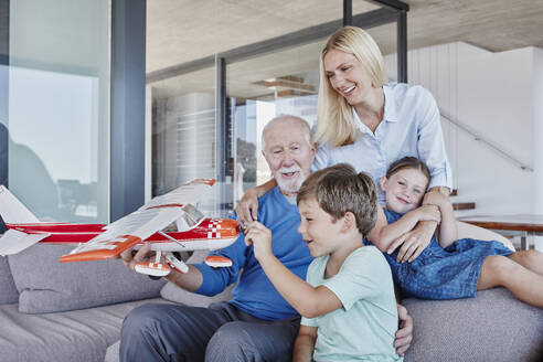 Älterer Mann, der ein Spielzeugflugzeug hält und mit einem Jungen spielt, während er neben seiner Tochter und einem Mädchen im Wohnzimmer sitzt - RORF02790