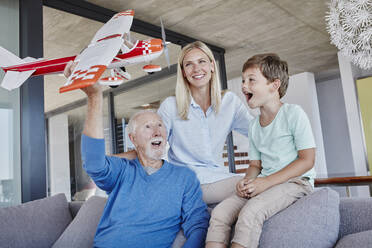 Senior man holding toy airplane playing with grandchild while sitting by woman in living room - RORF02791