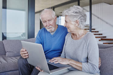 Senior couple with laptop sitting on sofa in living room - RORF02792