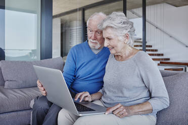 Surprised man looking at laptop used by woman while sitting in living room - RORF02794