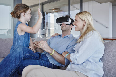 Mädchen winkt Vater zu, der einen Virtual-Reality-Simulator trägt, der bei einer Frau im Wohnzimmer sitzt - RORF02799