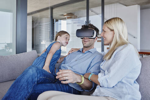 Lächelnder Mann, der einen Virtual-Reality-Simulator trägt, sitzt mit Frau und Tochter im Wohnzimmer - RORF02800