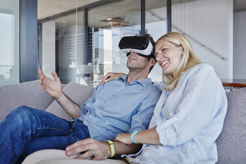 Lächelnde Frau sitzt neben einem Mann, der einen Virtual-Reality-Simulator im Wohnzimmer trägt - RORF02801