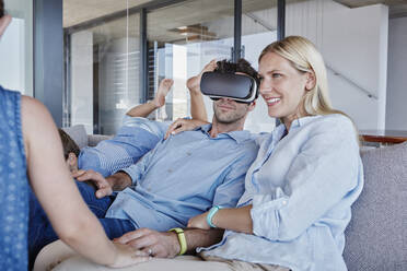 Man wearing virtual reality simulator sitting by woman while children playing in living room - RORF02802