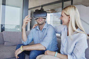 Cheerful blond woman looking at man wearing virtual reality simulator sitting in living room - RORF02803