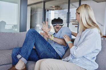 Excited man wearing virtual reality simulator sitting by cheerful woman in living room - RORF02804