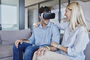 Cheerful woman with arm around looking at man wearing virtual reality simulator in living room - RORF02805