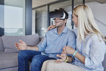 Blonde Frau, die den Arm um einen fröhlichen Mann legt, der einen Virtual-Reality-Simulator im Wohnzimmer trägt - RORF02806