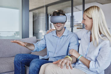 Cheerful woman looking at man wearing virtual reality simulator gesturing while sitting in living room - RORF02807