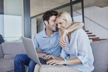Lächelnder Mann umarmt Frau, die einen Laptop benutzt, während sie auf dem Sofa im Wohnzimmer sitzt - RORF02811