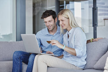 Smiling couple using laptop while doing online shopping sitting in living room at home - RORF02812
