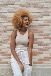 Happy woman with Afro hairstyle looking away while leaning on brick wall - AGOF00114