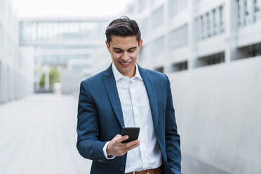 Lächelnder männlicher Berufstätiger, der im Freien stehend eine Textnachricht über sein Smartphone verschickt - DIGF15680