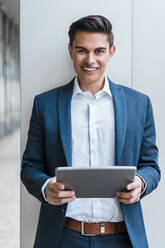 Lächelnder männlicher Fachmann, der ein digitales Tablet vor einer Wand im Korridor hält - DIGF15683