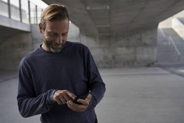 Smiling male professional text messaging through smart phone while standing under bridge - AUF00685