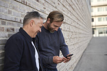 Cheerful male professional looking at friend using smart phone by wall - AUF00696