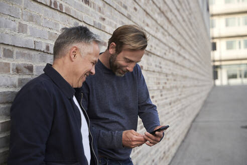 Fröhlicher männlicher Berufstätiger, der einen Freund ansieht, der sein Smartphone an der Wand benutzt - AUF00696