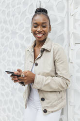 Lächelnde Frau mit Smartphone vor einer weißen Wand stehend - JRVF00896