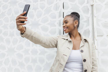 Smiling woman taking selfie through smart phone while standing in front of white wall - JRVF00897