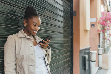 Schöne lächelnde Frau, die ein Smartphone benutzt, während sie vor einem Fensterladen steht - JRVF00900