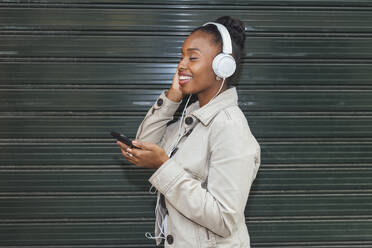 Smiling woman with eyes closed listening music through headphones in front of shutter - JRVF00901