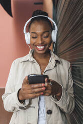 Glückliche Frau mit Kopfhörern, die eine Textnachricht über ihr Mobiltelefon sendet, während sie Musik hört, die von einem Fensterladen kommt - JRVF00903