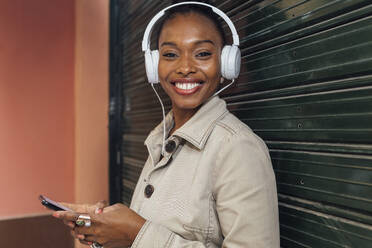 Smiling woman listening music through headphones while standing with mobile phone in front of shutter - JRVF00905