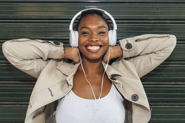 Cheerful woman wearing headphones while listening music in front of shutter - JRVF00906