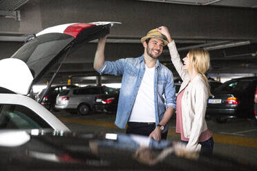 Young woman putting trilby on boyfriend's head in airport parking lot - AUF00717