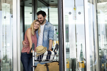 Man embracing girlfriend while standing with luggage at airport terminal - AUF00727