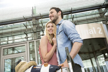 Smiling woman embracing boyfriend while arriving with luggage trolley from airport terminal - AUF00728