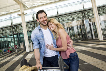 Happy young couple with luggage trolley standing at airport terminal - AUF00729