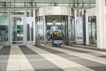 Young man with luggage trolley at airport terminal - AUF00732