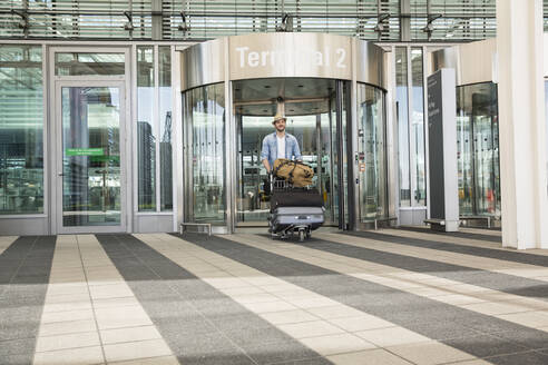 Young man with luggage trolley at airport terminal - AUF00732