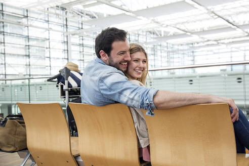 Lächelndes junges Paar wartet auf Sitze im Abflugbereich des Flughafens - AUF00737
