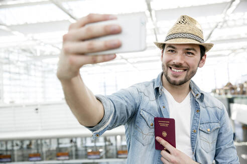 Junger Mann hält seinen Reisepass und macht ein Selfie im Abflugbereich eines Flughafens - AUF00739