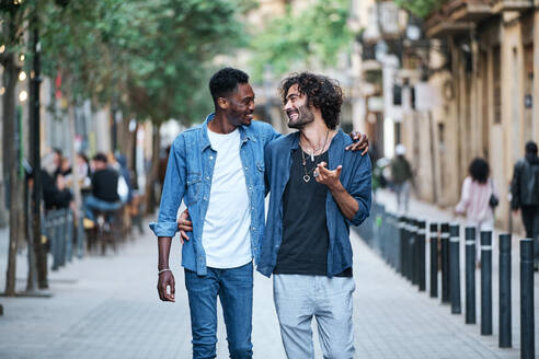 Gay man with arm around talking to friend while walking on footpath - AGOF00135