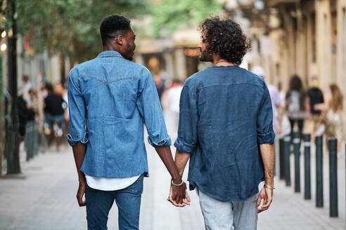 Gay couple holding hands looking at each other while walking on footpath - AGOF00137